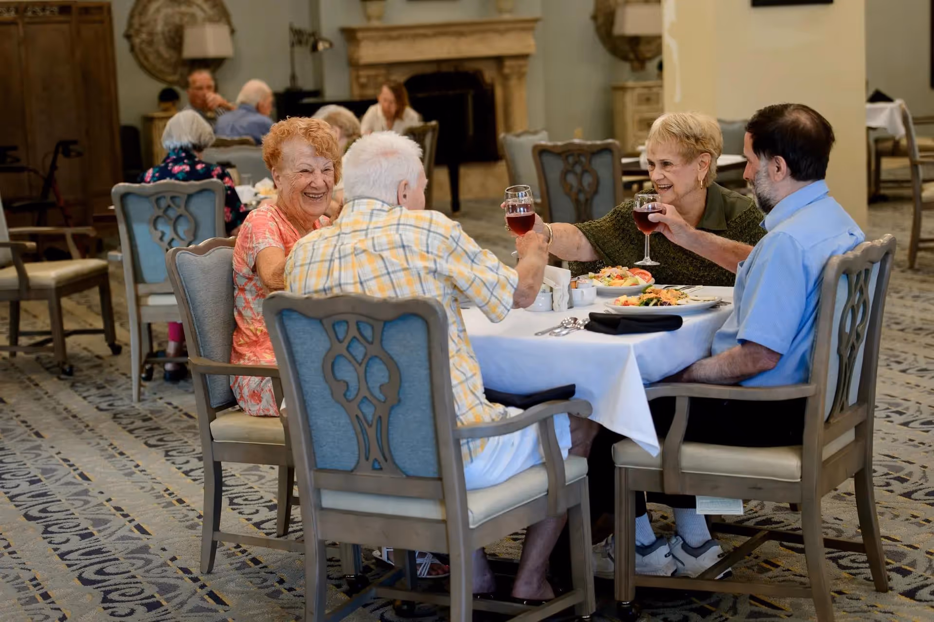 Four elderly people sitting around a dining table in a senior living facility, smiling and toasting with glasses of red wine. The table is set with plates of food and silverware. The room has a patterned carpet, upholstered chairs, and a fireplace in the background.