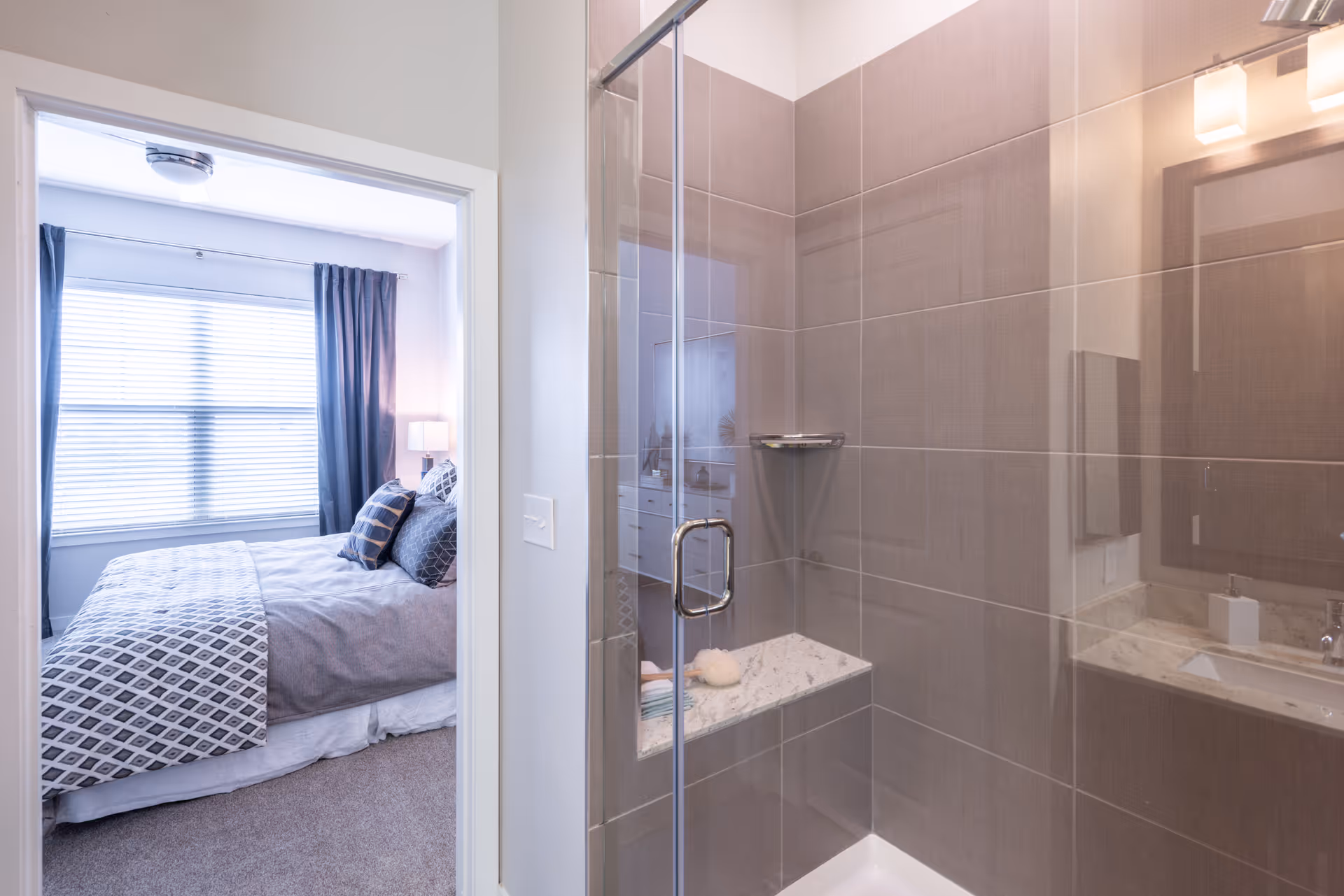 A modern bathroom with a glass-enclosed shower and tiled walls, visible next to a bedroom with a bed and window.