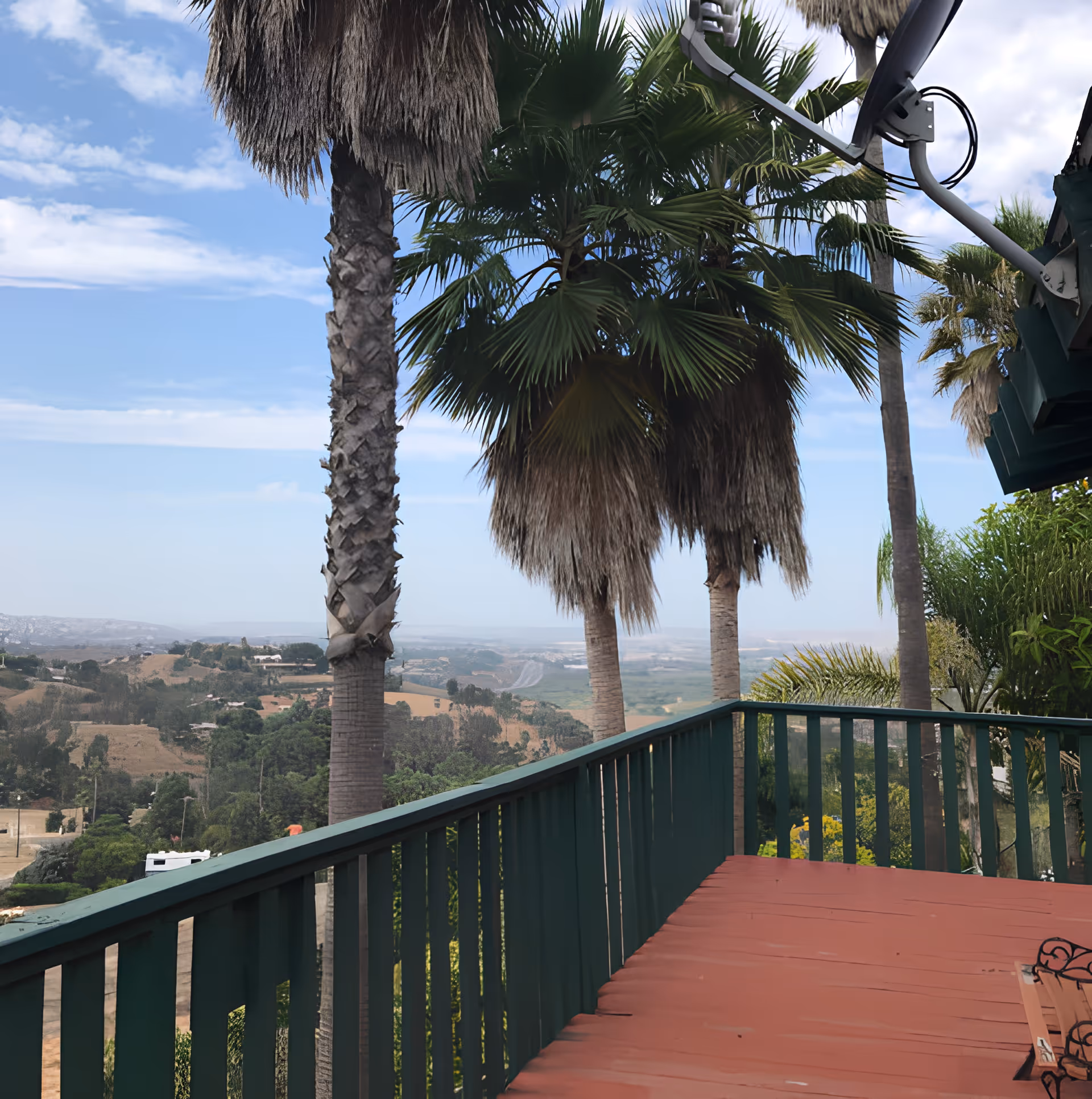 View from a wooden deck with green railings overlooking a landscape with palm trees, hills, and a partly cloudy sky.