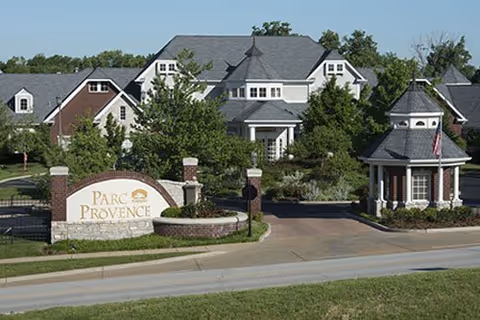 Entrance to Parc Provence with a stone sign, gated driveway, guardhouse, and residential buildings behind it.
