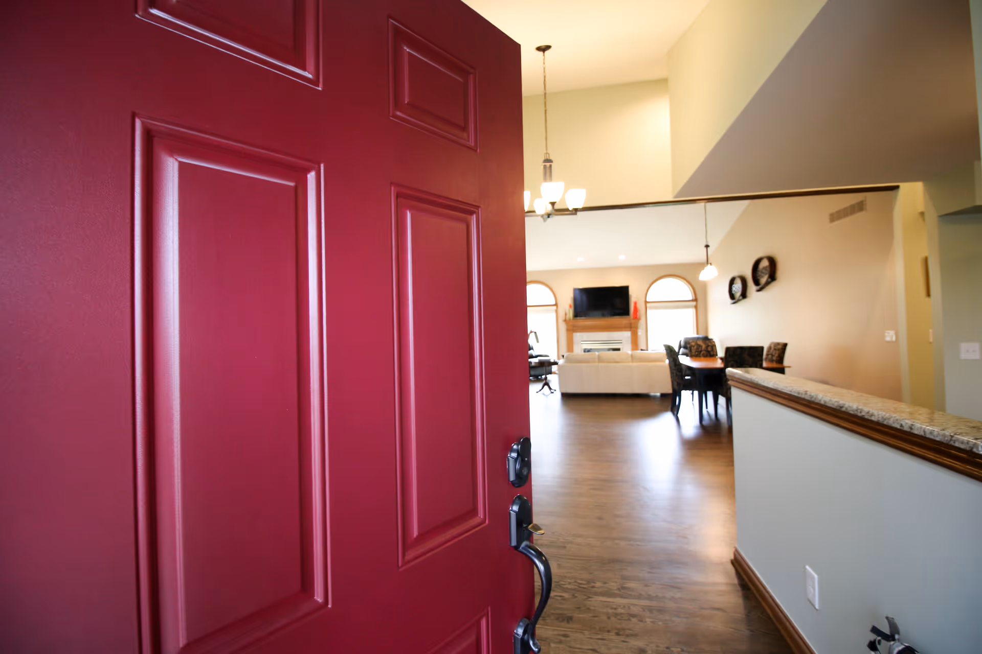 View through a partially open red front door into a spacious living area with hardwood floors, a white sofa, a dining table with chairs, a fireplace with a mounted TV above it, and large windows letting in natural light.
