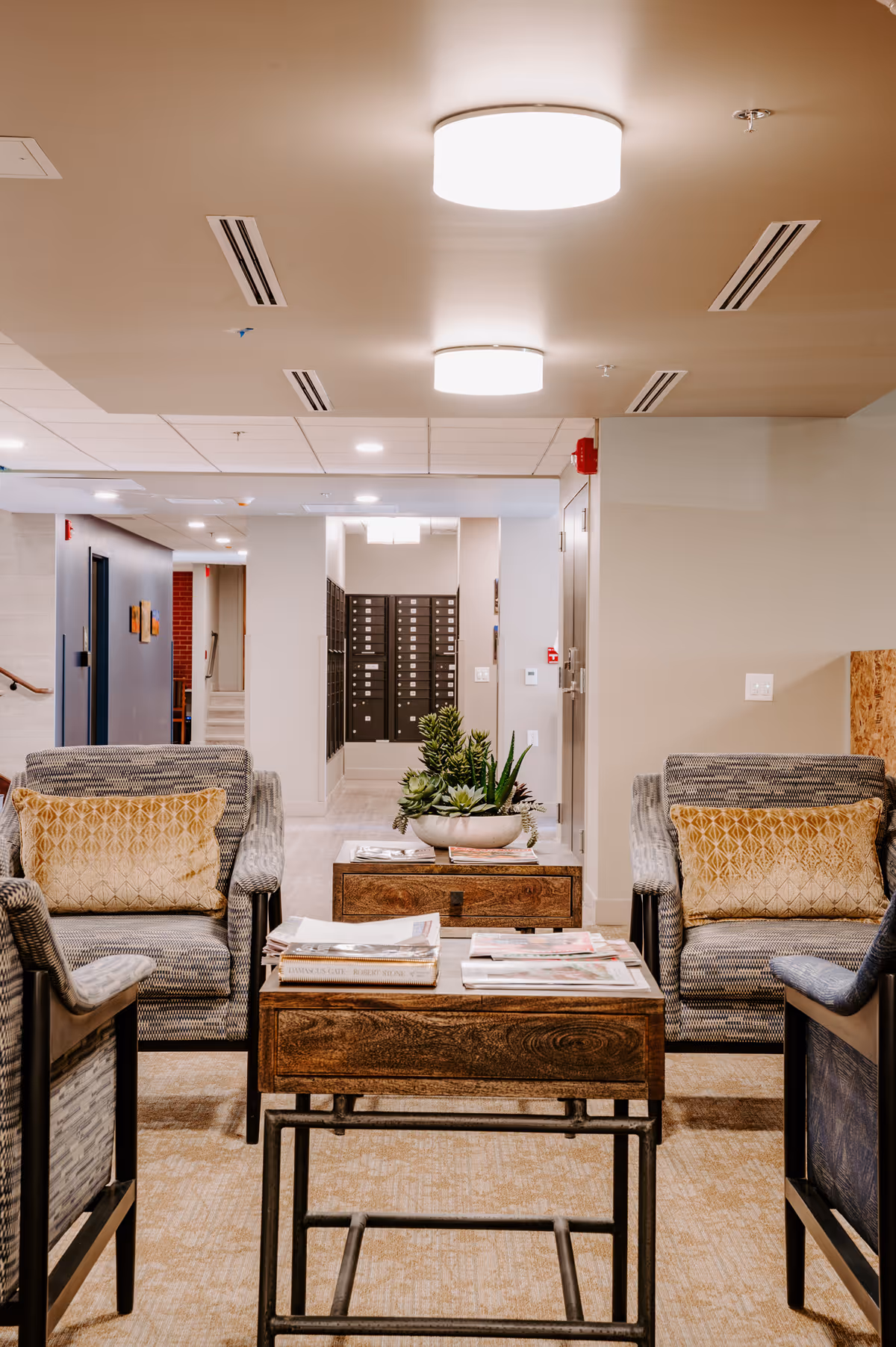 A cozy seating area in a senior living facility with four cushioned armchairs arranged around two wooden coffee tables. The tables have magazines and a decorative plant. The background shows a hallway with mailboxes and neutral-colored walls and ceiling lights.