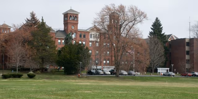 Front view of a large red-brick senior living building with towers, leafless trees, a grassy lawn, and parked cars.