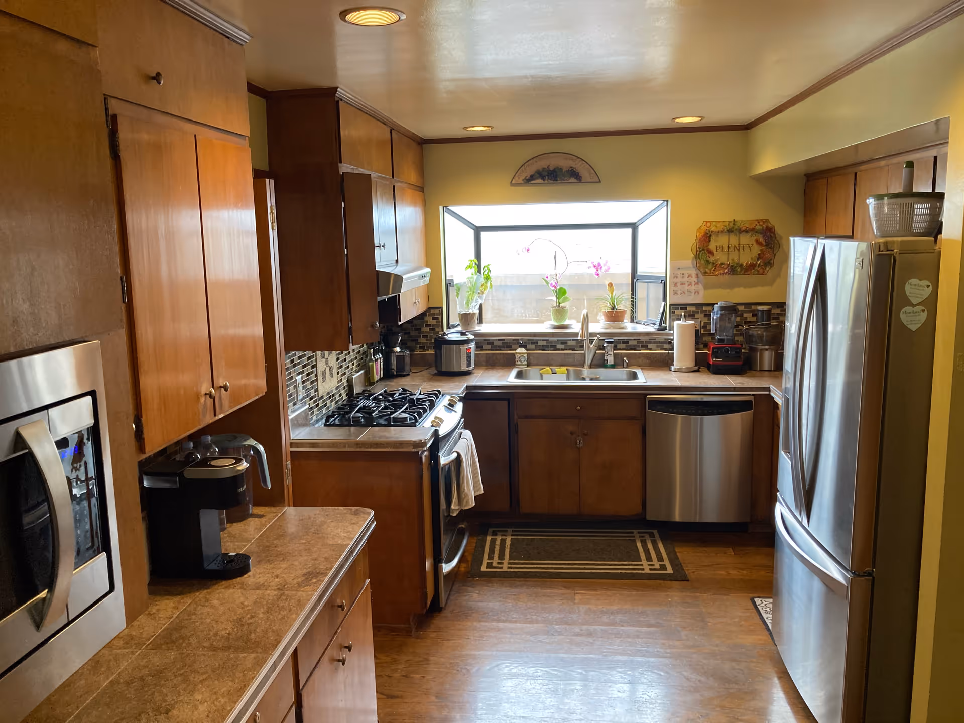 A kitchen with wooden cabinets, a stainless steel refrigerator, dishwasher, microwave, and gas stove. There is a coffee maker on the counter, a window above the sink with potted plants on the windowsill, and various kitchen appliances on the countertops.