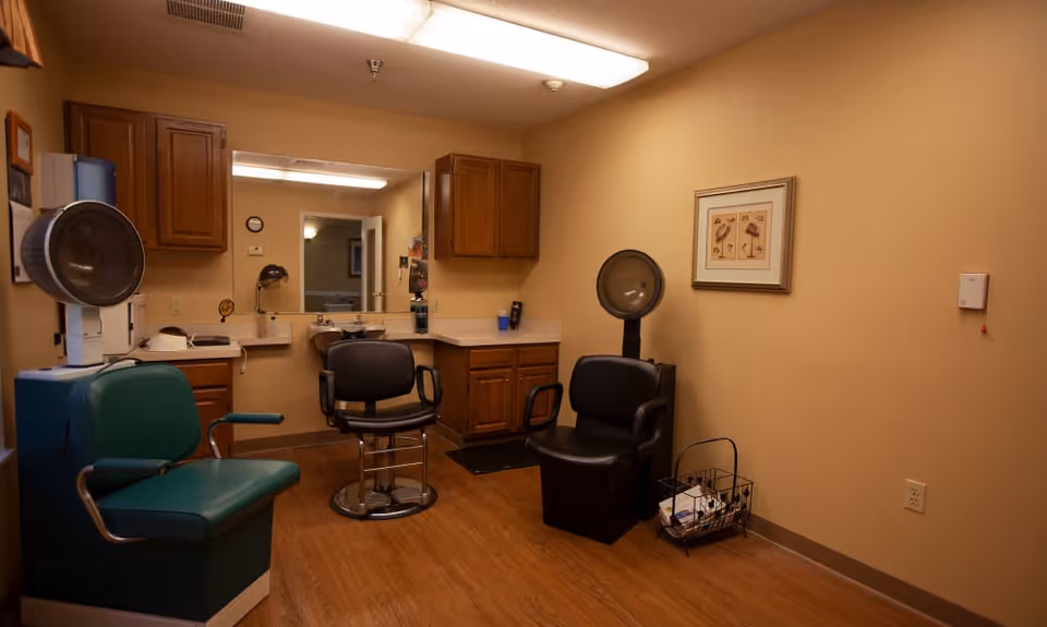 Small salon room with hair-styling chairs, hooded dryers, a large mirror and wooden cabinets.