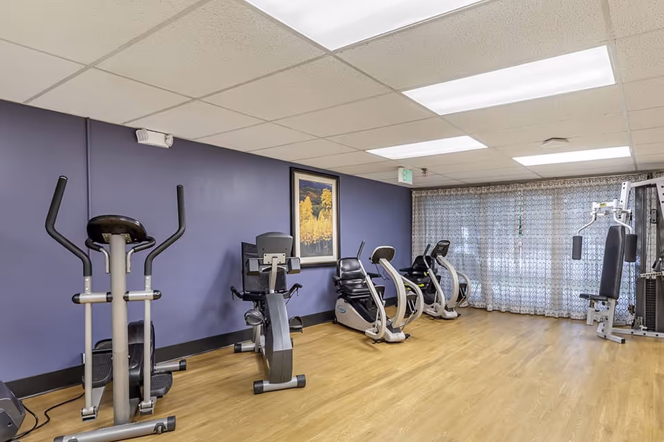 Small fitness room with several cardio machines and a weight station against a purple wall and wood floor.