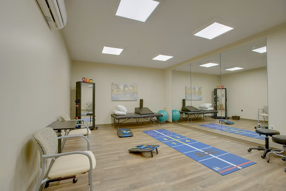 Bright therapy/exercise room with a mirrored wall, treatment tables, exercise mats, chairs and fitness equipment.