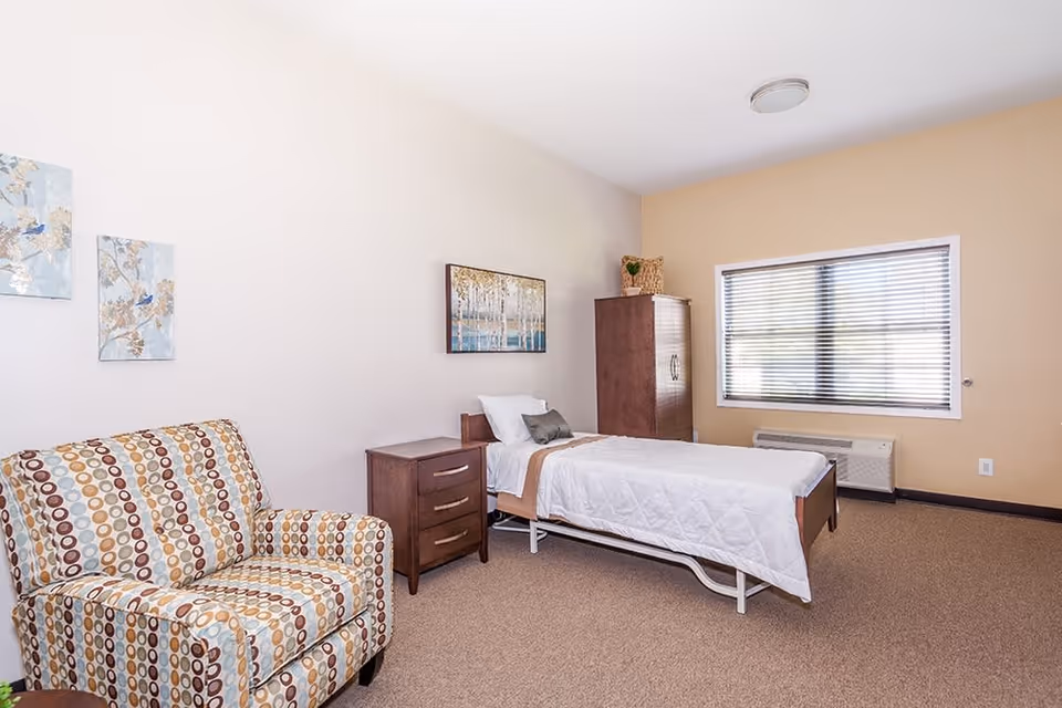 A bright and clean bedroom in an assisted living facility featuring a single bed with white bedding, a wooden nightstand with drawers, a tall wooden wardrobe, a patterned armchair with circular designs, and a window with blinds allowing natural light to enter the room. The walls are decorated with framed artwork.