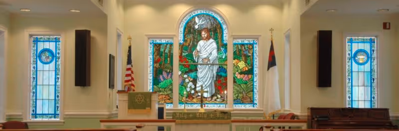 Interior chapel with a large central stained-glass window depicting Jesus, flanked by tall stained-glass panels, an altar, flags, and pews.