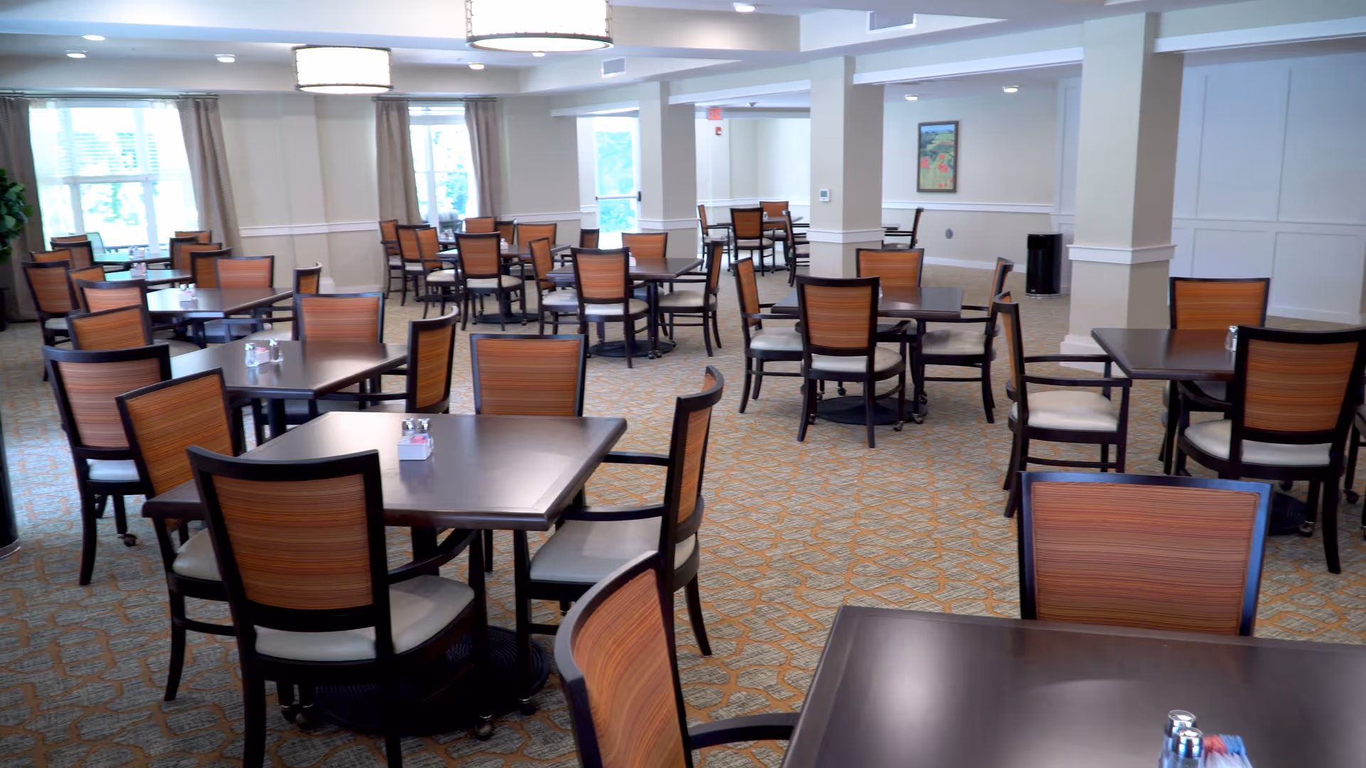 A spacious dining room with multiple dark wood tables and chairs with wooden backs and light cushioned seats. The room has beige walls, patterned carpet, large windows with curtains, and ceiling lights.