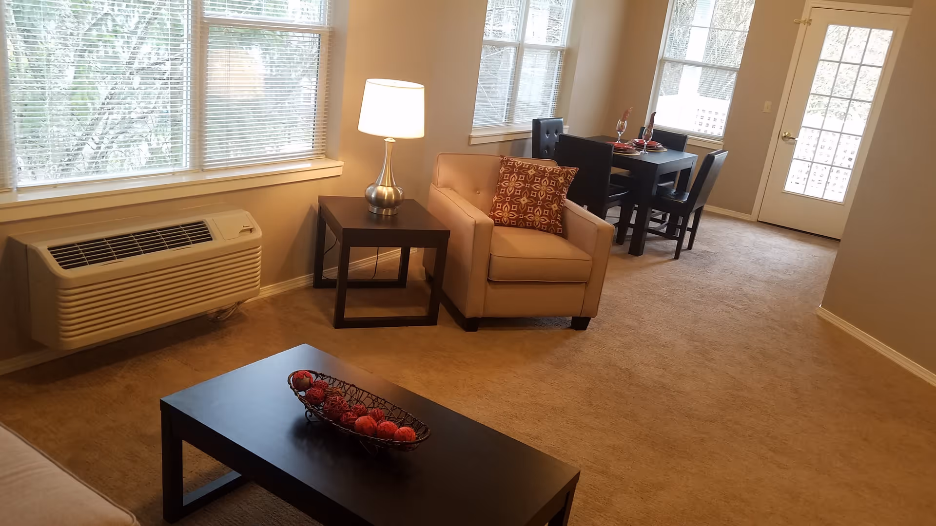 A cozy living area with beige carpet, a beige armchair with a patterned cushion, a dark wooden side table with a silver lamp, and a dark wooden coffee table with a decorative bowl of red spheres. In the background, there is a small dining table set with four chairs and place settings near a door with glass panes. Large windows with blinds let in natural light.