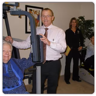 An elderly woman using exercise equipment in a room, assisted by a man in a white shirt and tie. A woman holding a folder stands in the background near another person seated on exercise equipment.