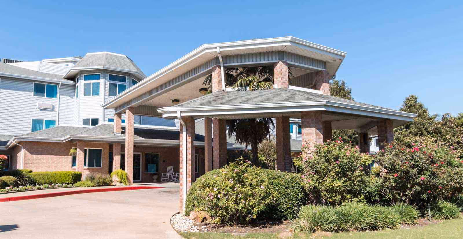 Front entrance of a multi-story senior living facility with a covered porte-cochere, brick columns, and landscaped shrubs.