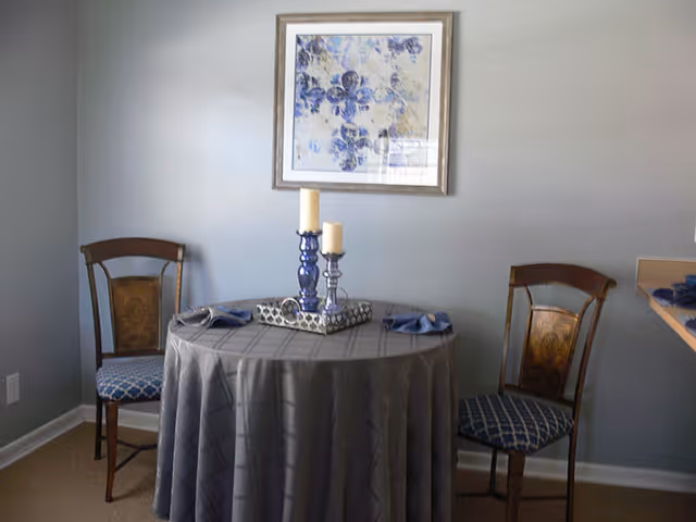 Small dining nook with a round table draped in a gray tablecloth, two wooden chairs, a candle centerpiece, and blue floral artwork on the wall.