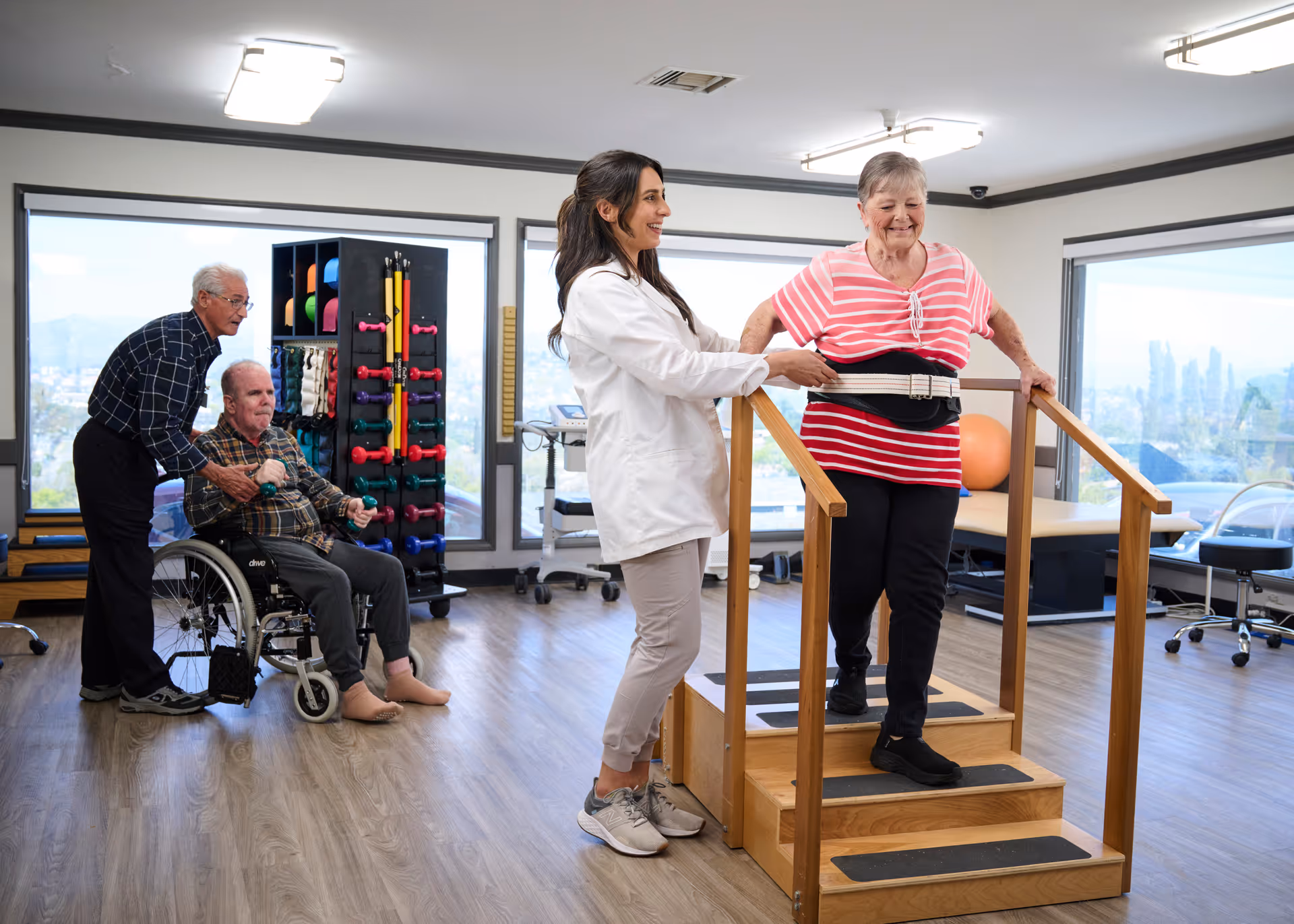 A physical therapy session in a rehabilitation room with large windows. An elderly woman wearing a pink striped shirt is walking up wooden steps with handrails, assisted by a female therapist in a white coat. In the background, an elderly man in a wheelchair is holding small dumbbells while another elderly man stands behind him, supporting him. The room has exercise equipment and a bright, spacious atmosphere.