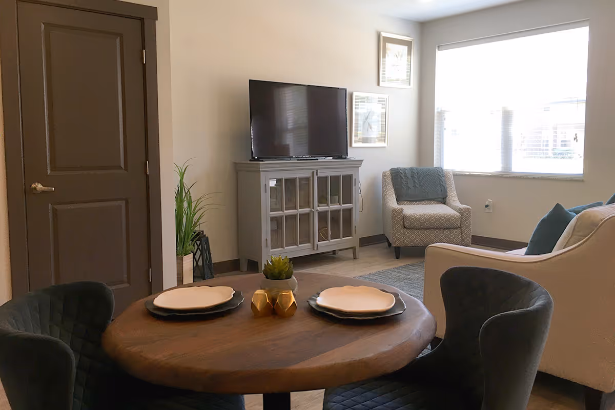 A cozy living room area featuring a round wooden dining table set with plates and decorative items, surrounded by four dark cushioned chairs. In the background, there is a TV on a gray cabinet, a patterned armchair with a blue throw, a beige sofa with blue pillows, and a large window letting in natural light. A closed brown door and a tall green plant are also visible.
