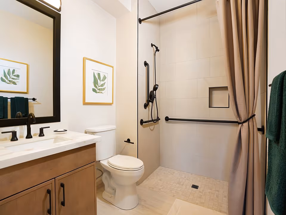 A modern bathroom featuring a white toilet, a wooden vanity with a white countertop and black faucet, a large mirror with a black frame, and a walk-in shower with beige tiles, a black handheld showerhead, grab bars, and a beige shower curtain tied back. A framed botanical print hangs on the wall above the toilet, and green towels are visible on a towel rack.