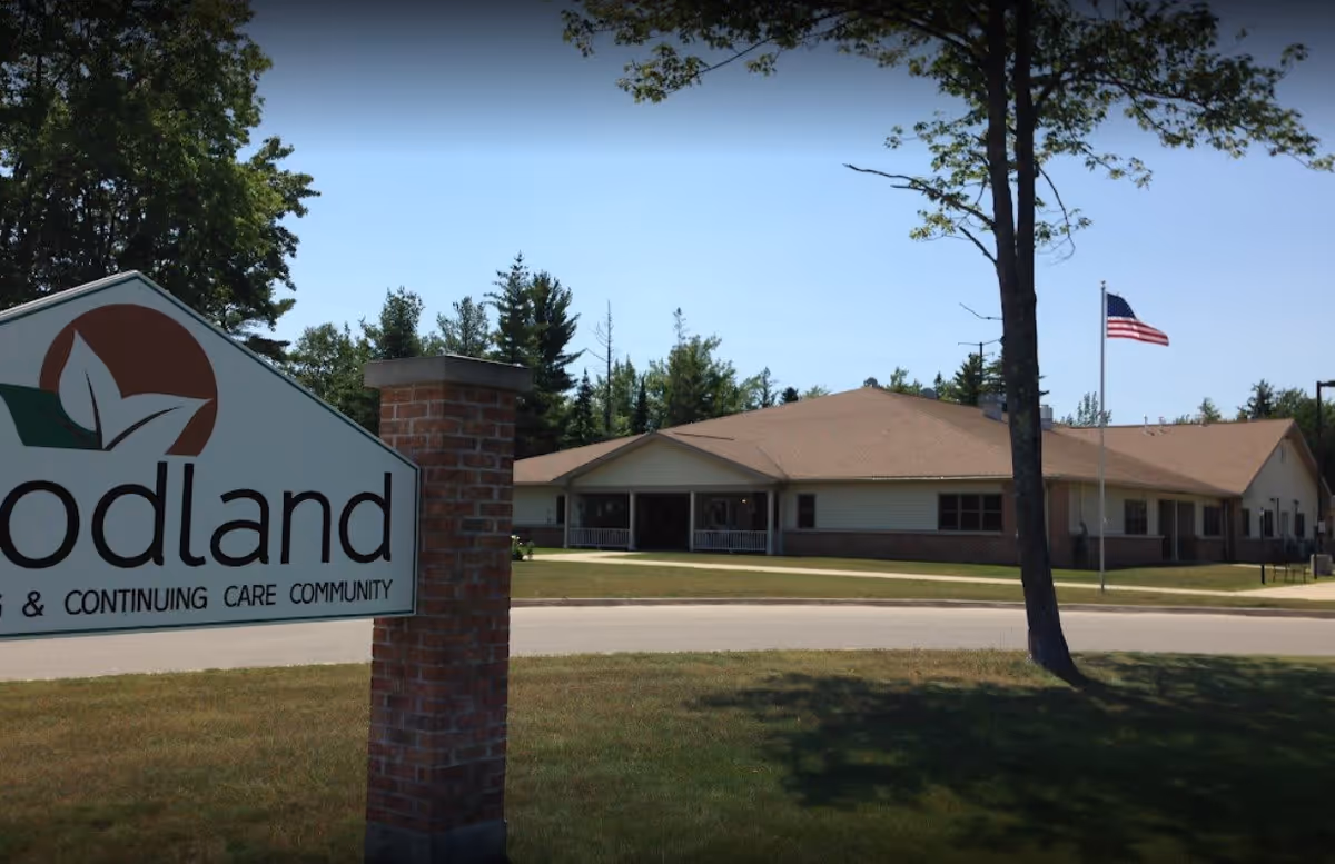 Exterior view of Woodland Assisted Living and Continuing Care Community building with a large sign in the foreground, a tree, and an American flag on a flagpole on a sunny day.