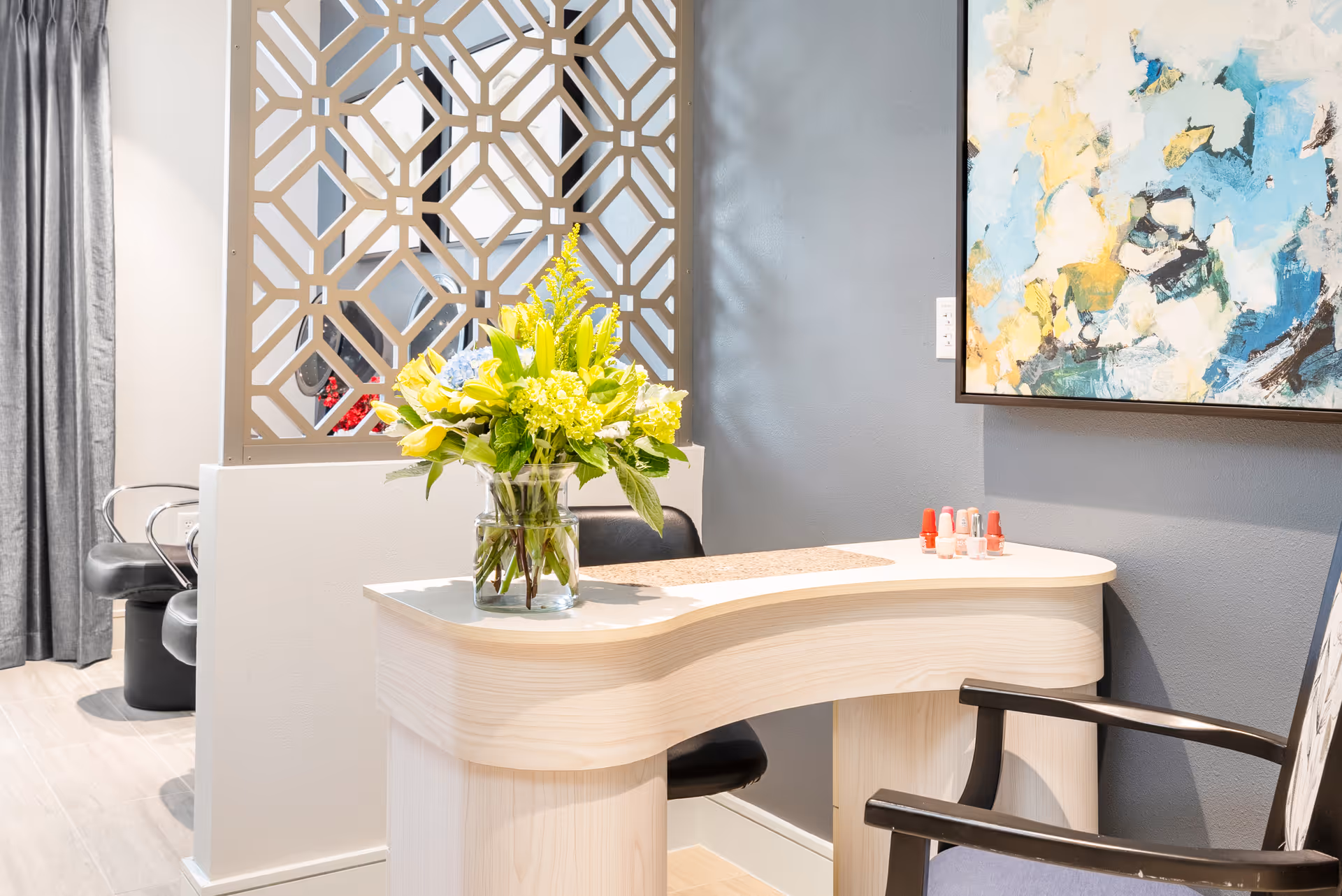 A small manicure station with a curved light wood table, a glass vase with a bouquet of yellow and green flowers, and several bottles of nail polish on the table. There is a black chair in front of the table and a decorative partition with a geometric pattern behind it. A colorful abstract painting hangs on the gray wall to the right.