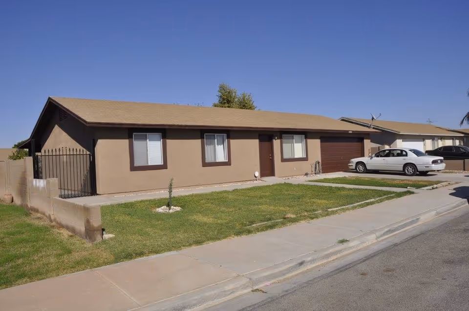 Single-story residential building with beige exterior walls and brown trim, featuring three windows and a brown front door. There is a small grassy front yard with a young tree and a concrete driveway with a white car parked on it. The sky is clear and blue.