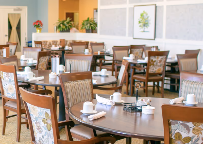 A bright and neatly arranged dining room with multiple round wooden tables and chairs. Each table is set with white cups, saucers, and rolled white napkins. The chairs have patterned upholstery on the backrests. The room has light-colored walls with framed artwork and decorative plants on a ledge in the background.