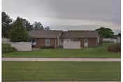 Single-story brick residential building with a garage in the center, surrounded by a grassy lawn and some trees under a cloudy sky.