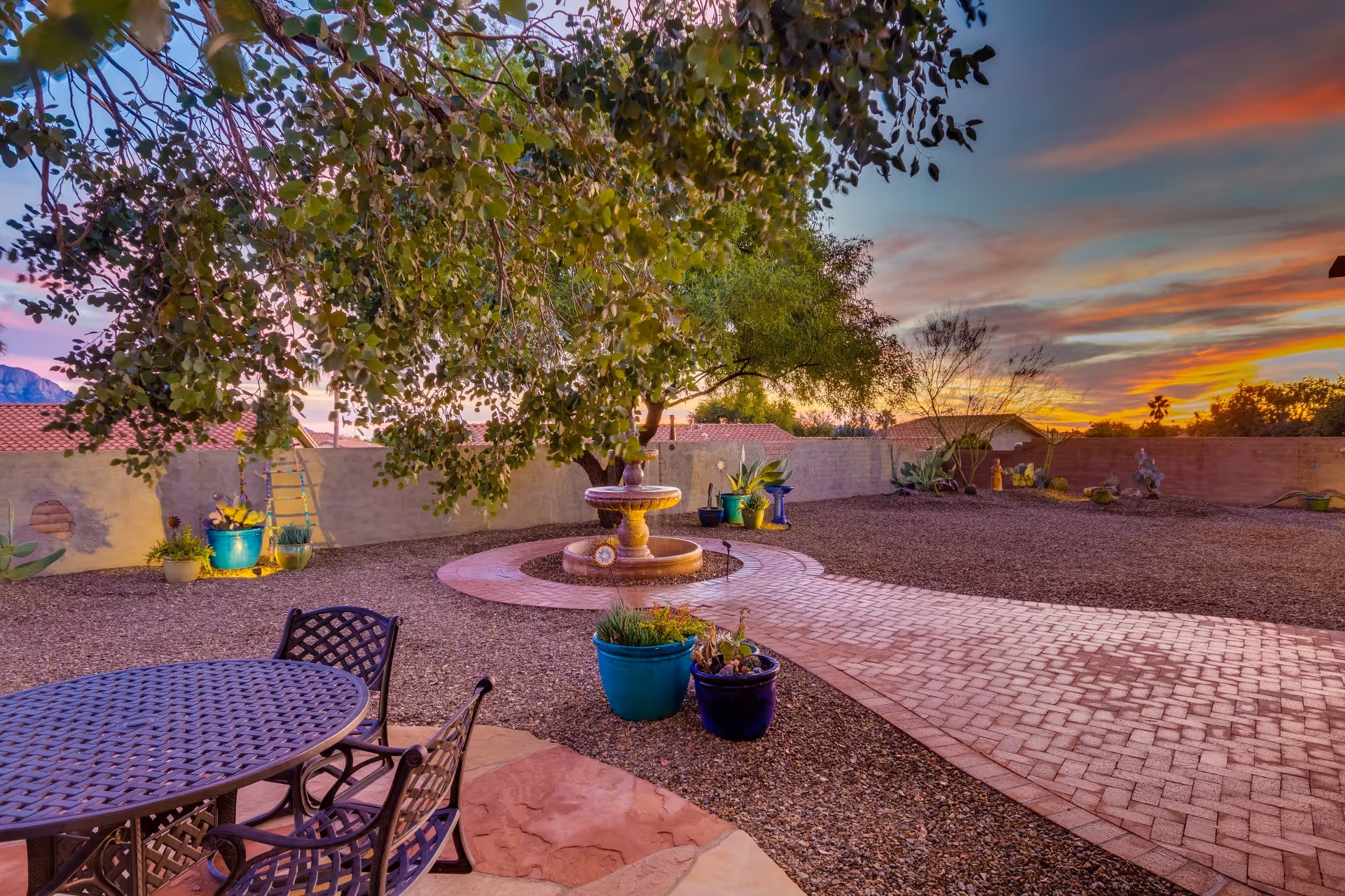 Landscaped backyard patio with a round metal table and chairs, potted plants, a central fountain, and a large tree at sunset.