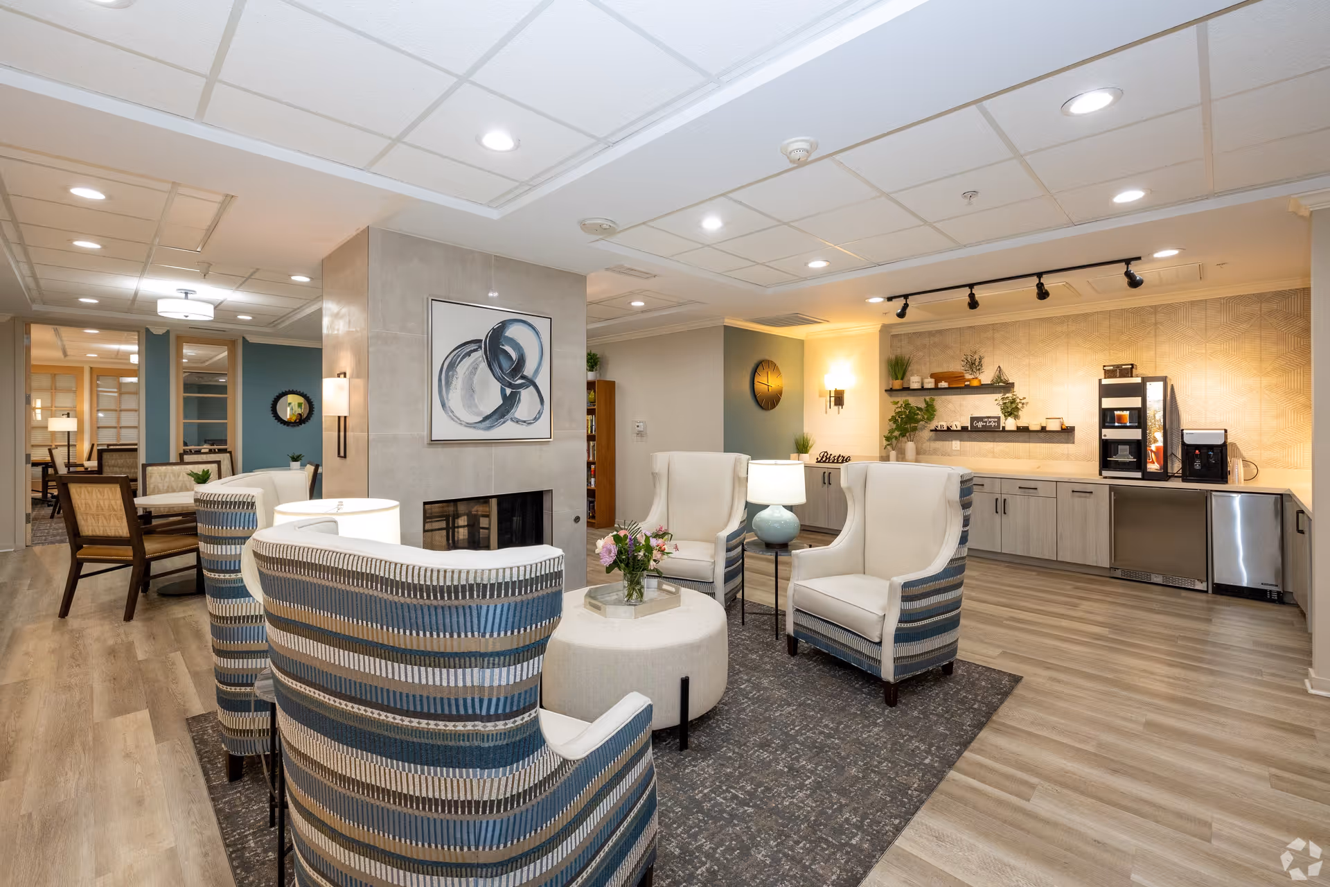 A cozy senior living facility common area with a seating arrangement of four striped armchairs around a round ottoman with a flower vase on top. The room features a fireplace with abstract art above it, a kitchenette area with a coffee machine, and modern lighting fixtures. The flooring is wood-style laminate, and the walls are decorated with plants and a clock.