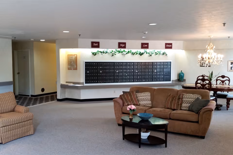 Interior view of a senior living facility lobby area with a brown couch and a striped armchair, a coffee table with a vase of flowers and a bowl, a dining table with chairs, a chandelier, and a wall of mailboxes decorated with green garland and signs that read Faith, Hope, Love, Dream.