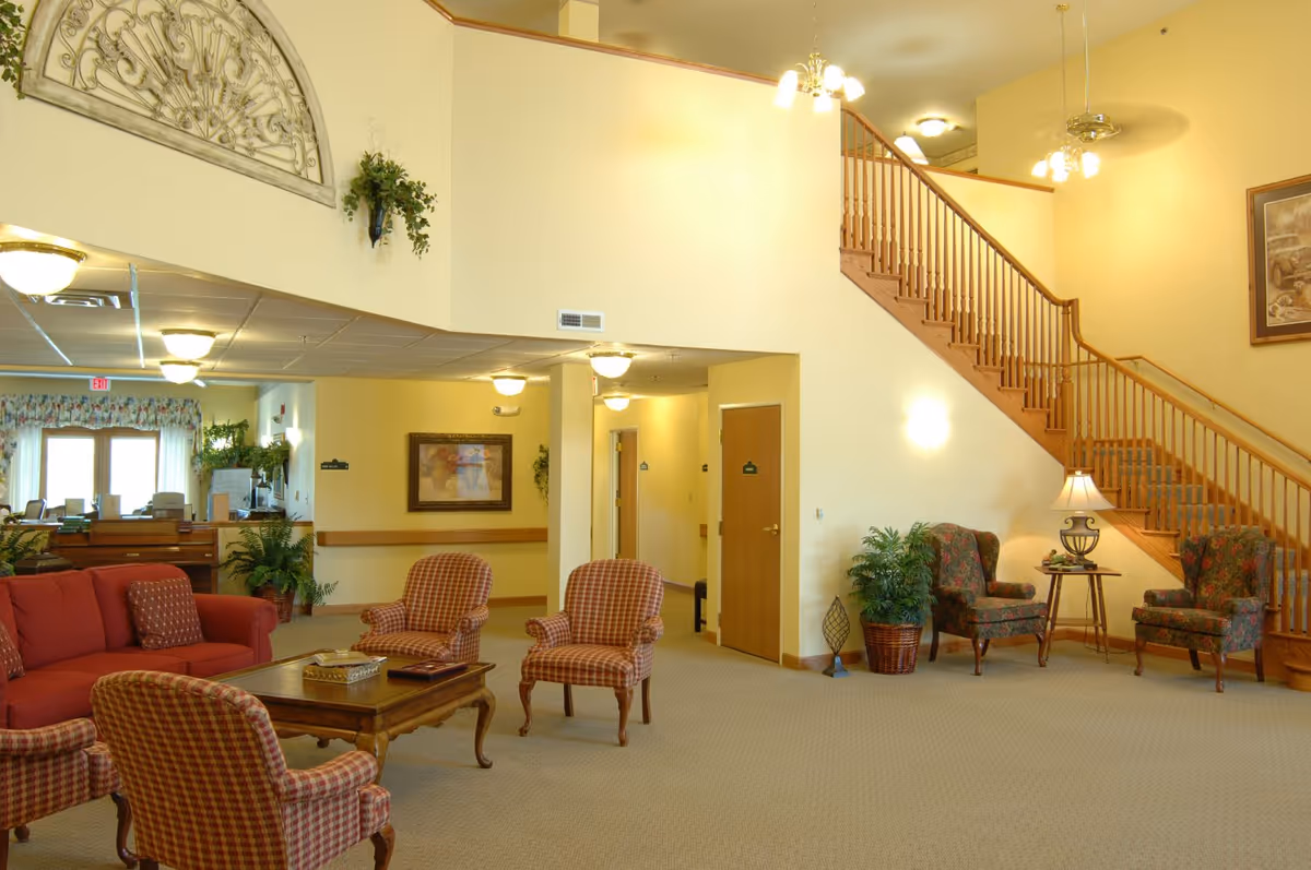 A spacious and well-lit senior living facility common area with a red sofa, four plaid armchairs around a wooden coffee table, two floral armchairs near a wooden staircase, potted plants, wall art, and ceiling lights.