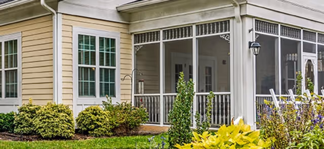 Exterior view of a building corner with beige siding and white trim, featuring large screened windows and a porch area. The foreground includes green shrubs, plants, and a well-maintained lawn.