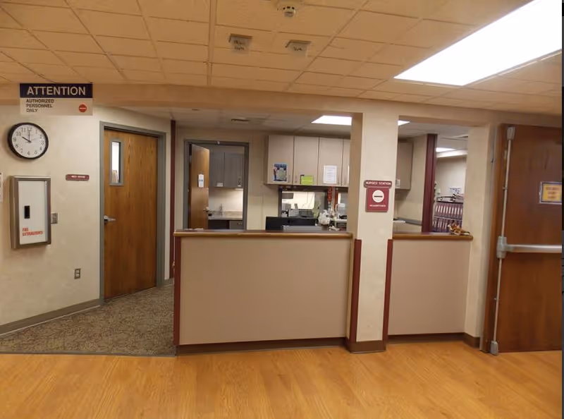 Interior view of a nurse's station in a senior living facility with a counter, cabinets, and office equipment. There are wooden doors, a wall clock showing 9:40, and signs indicating 'Attention Authorized Personnel Only' and 'Nurses' Station No Admittance'. The floor is a combination of wood and carpet.