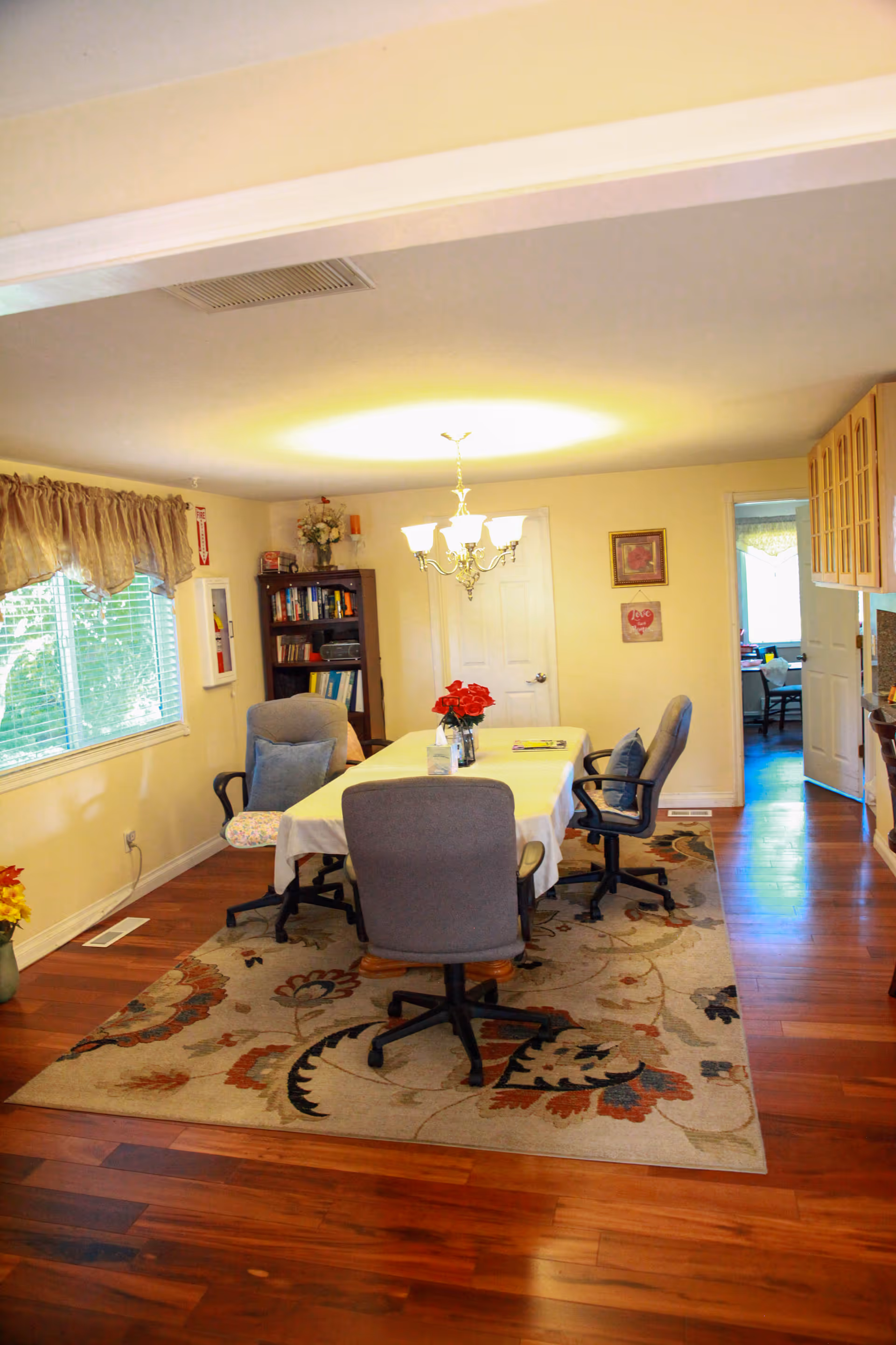 A dining room with a rectangular table covered with a white tablecloth, surrounded by four office-style chairs on a patterned area rug. A chandelier hangs above the table, and a vase with red flowers is placed on it. There is a window with curtains on the left wall, a bookshelf with books and decorations in the corner, and a doorway leading to another room on the right. The floor is wooden, and the walls are painted light yellow.