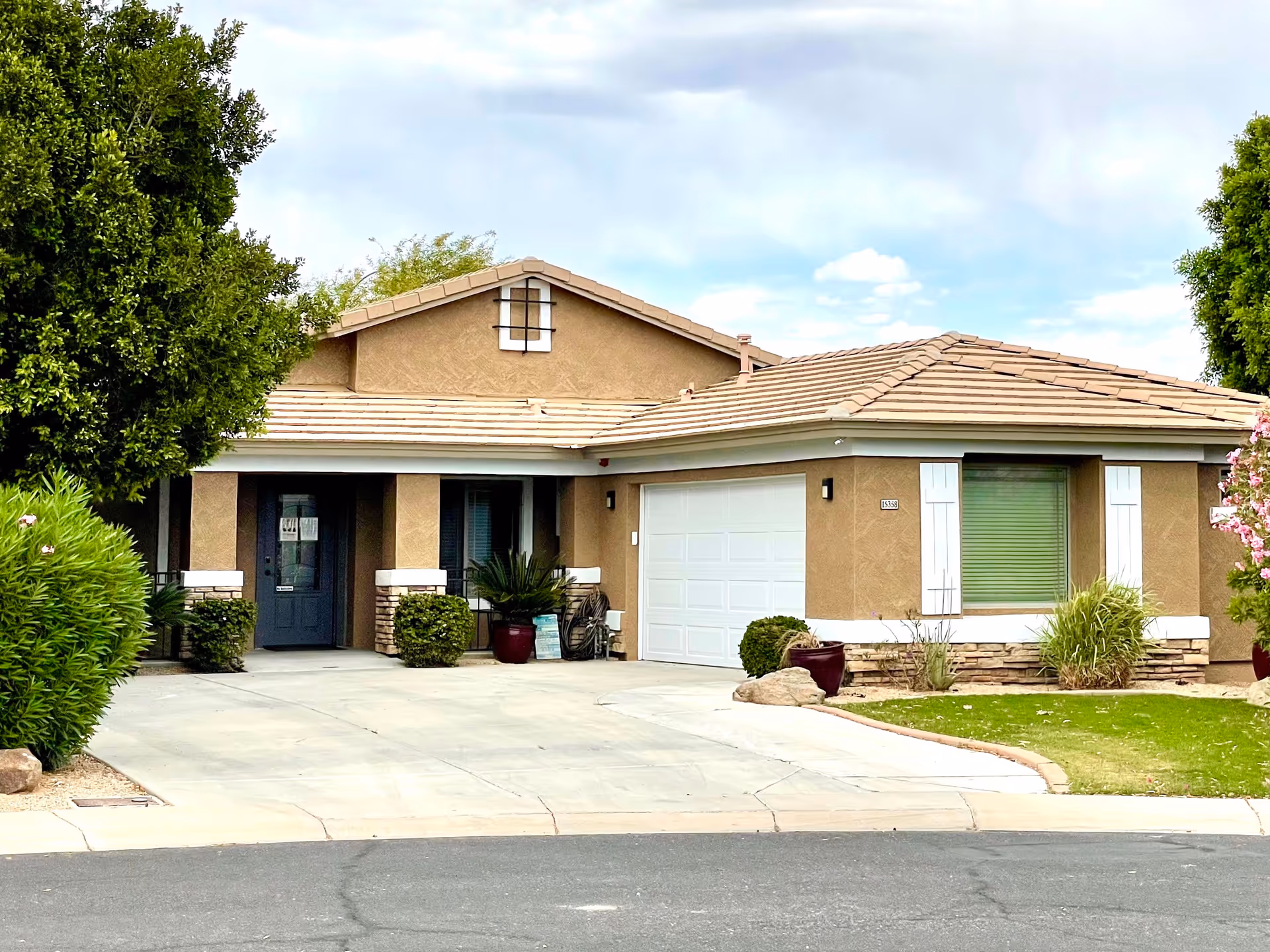 Front exterior view of a single-story residential building with a beige stucco finish, a tiled roof, a white garage door, and a dark front door. The driveway is concrete and there are green bushes and trees around the house, along with a small lawn area.
