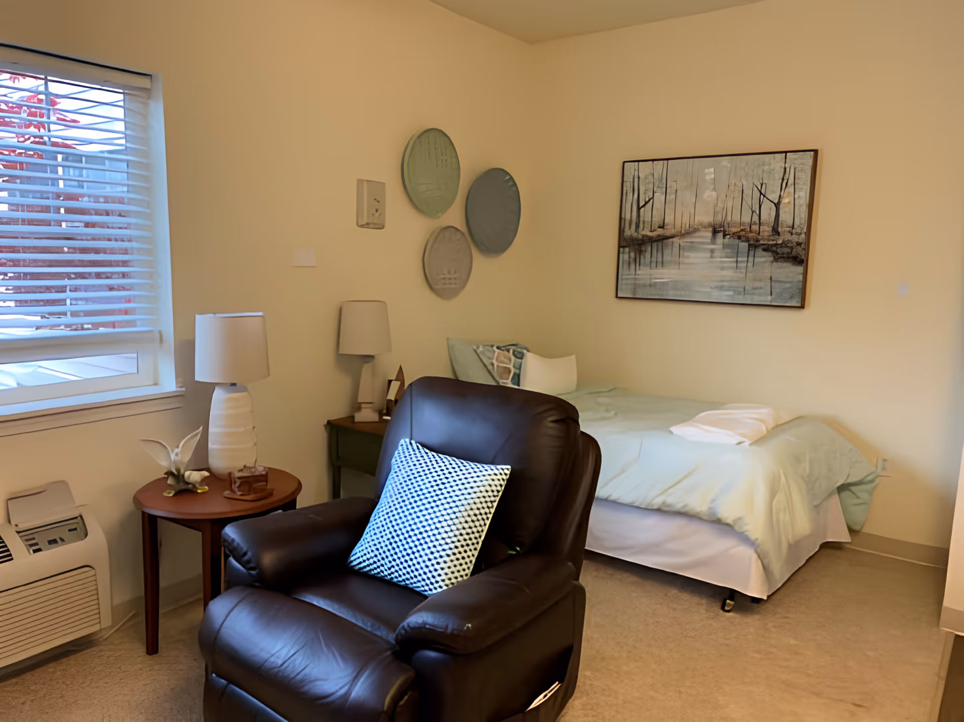 A cozy bedroom in a senior living facility with a single bed covered in light green bedding, a black leather recliner chair with a patterned pillow, a small round wooden side table with a white lamp and decorative items, a window with white blinds, and wall decorations including three round plates and a framed painting of a forest scene.