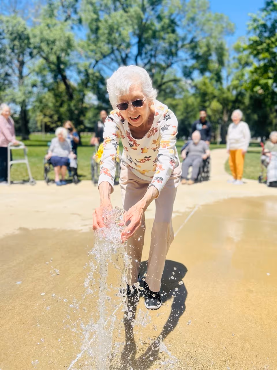 An elderly woman wearing sunglasses and a floral long-sleeve shirt is playing with water from a ground fountain in an outdoor park area. Several other elderly individuals, some seated and some standing, are visible in the background along with green trees and a clear blue sky.
