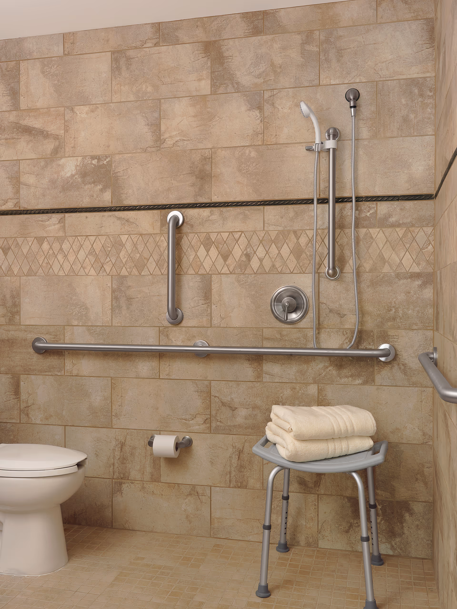 Accessible bathroom with beige tiled walls and floor, featuring a white toilet, multiple stainless steel grab bars, a handheld showerhead with adjustable height, and a gray shower chair with folded towels on top.