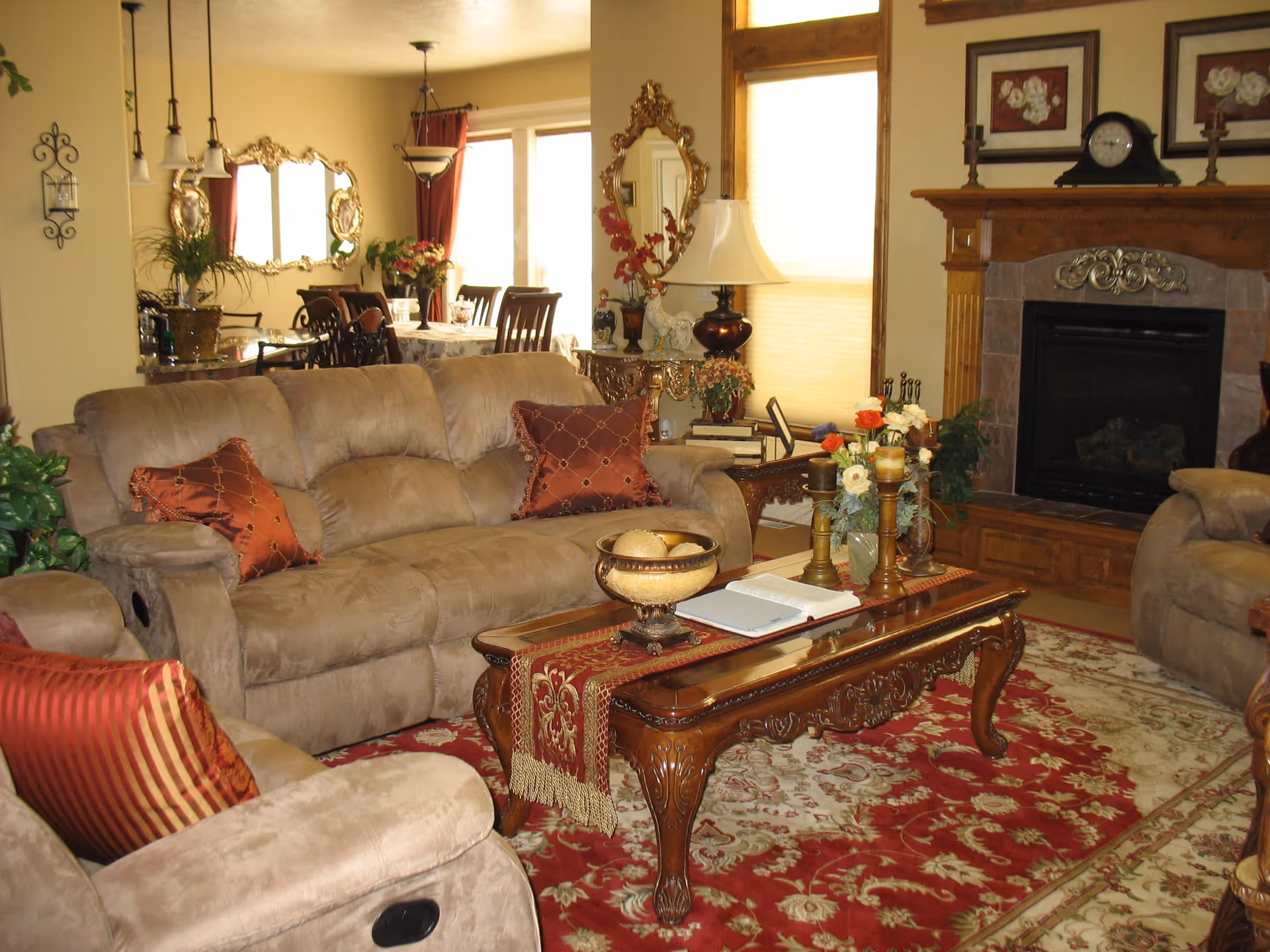 A cozy living room with beige plush sofas adorned with red and gold decorative pillows. A wooden coffee table with ornate carvings sits on a red and beige patterned rug, holding candles, a bowl, and an open book. In the background, there is a wooden fireplace with a clock and framed pictures above it, and a dining area with a table and chairs, mirrors, and plants.