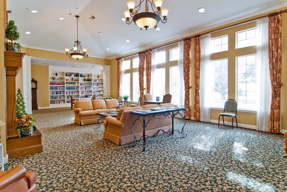 A spacious and well-lit living room area with large windows covered by patterned curtains allowing natural light to fill the room. The room features comfortable beige sofas arranged around a coffee table, a decorative fireplace with plants and flowers, and a bookshelf filled with books in the background. The carpet has an intricate floral pattern, and there are chandeliers hanging from the ceiling.