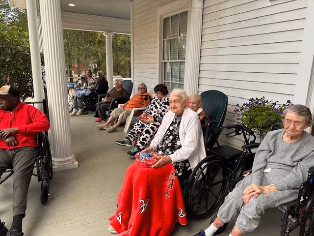 A group of elderly individuals sitting on a covered porch outside a white building. Some are in wheelchairs, others in chairs, and they appear to be enjoying the outdoor setting with greenery visible in the background.