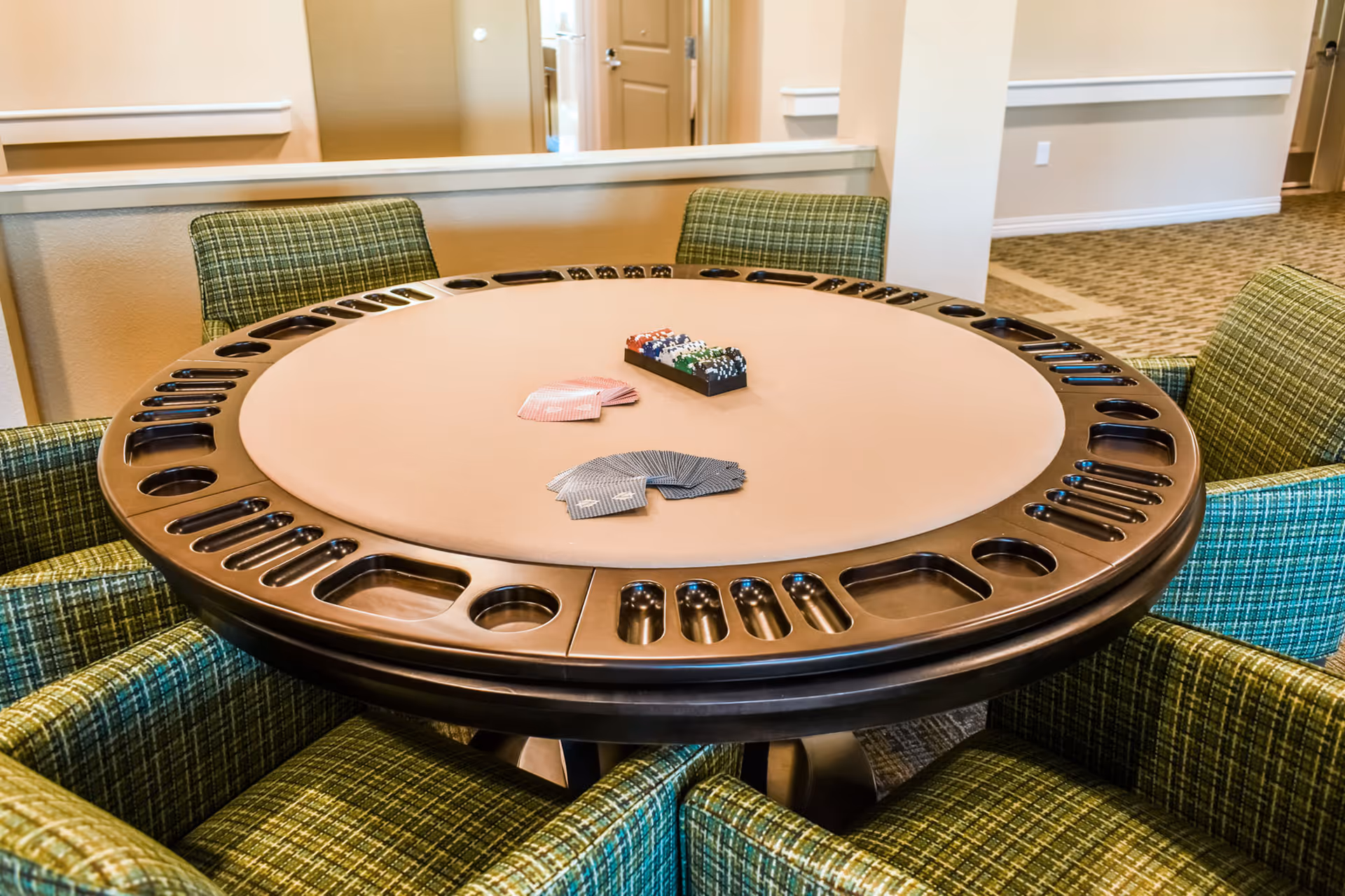 Round poker table with built-in cup holders and slots for poker chips, surrounded by green plaid upholstered chairs in a well-lit room with beige walls and carpeted floor.