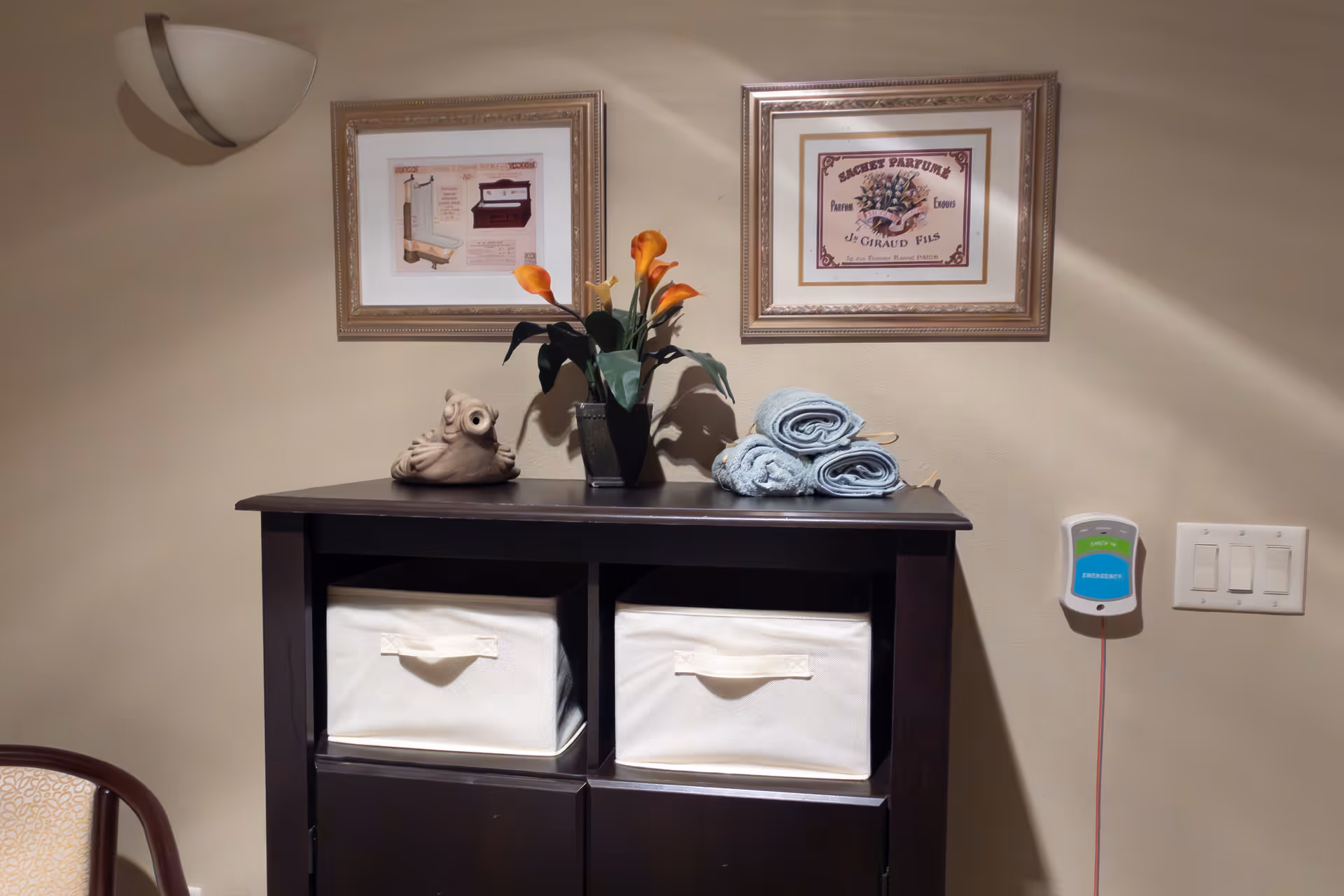 A dark wooden cabinet with two white fabric storage bins on the lower shelves. On top of the cabinet are three rolled light blue towels, a small owl figurine, and a vase with orange calla lilies. Two framed pictures hang on the beige wall above the cabinet. To the right of the cabinet, there is an emergency call button and a light switch panel. A wall-mounted light fixture is visible on the upper left corner.
