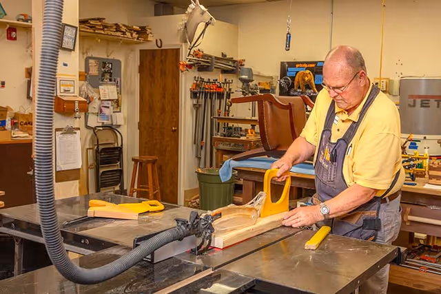 An elderly man wearing glasses and a yellow shirt with overalls is working in a woodworking shop. He is using a push block to guide a piece of wood through a table saw. The workshop is equipped with various woodworking tools and equipment, including clamps, a stool, and a mounted horse head decoration on the wall.