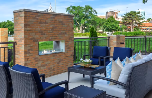 Outdoor patio seating area with wicker chairs and blue cushions around a low table in front of a brick fireplace, overlooking greenery and distant buildings.
