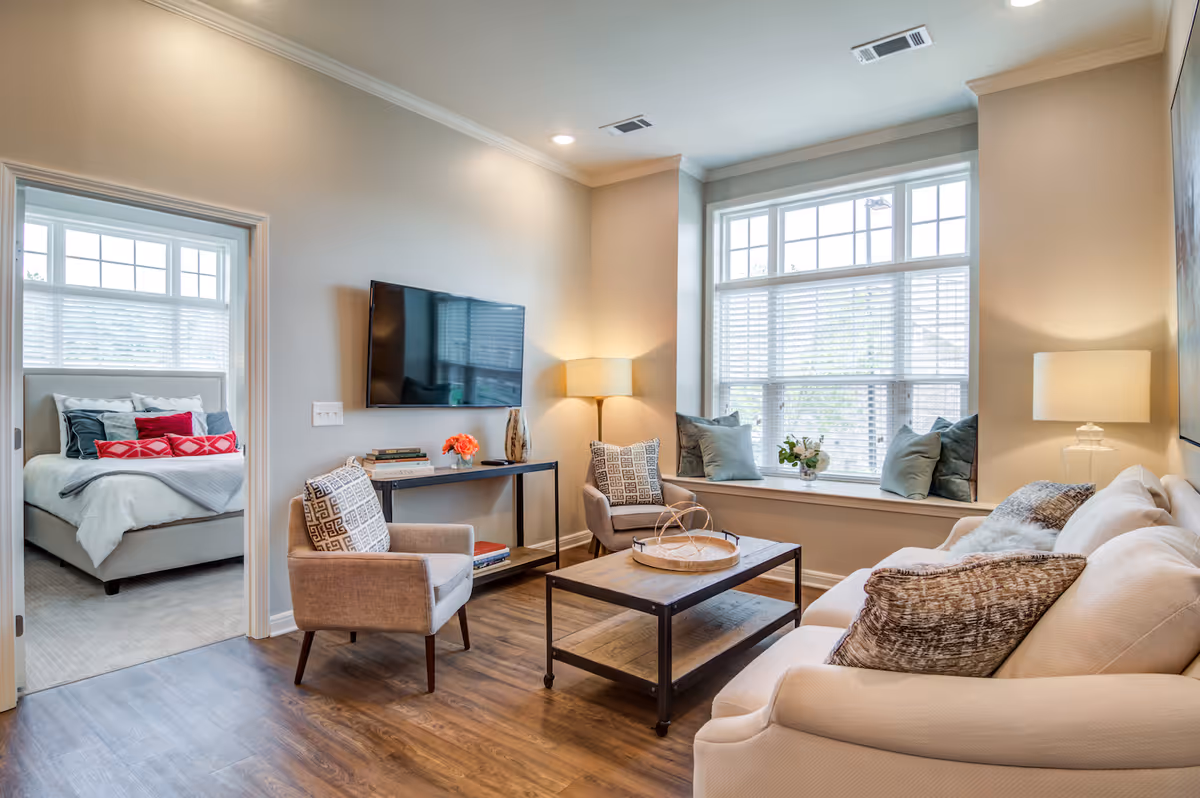 A cozy living room with a beige sofa, two armchairs with patterned pillows, a wooden coffee table, and a large window with cushions on the window seat. A flat-screen TV is mounted on the wall above a console table with books and decorative items. The room has wooden flooring and soft lighting from a floor lamp and a table lamp. Through an open doorway, a bedroom with a neatly made bed and multiple pillows is visible.