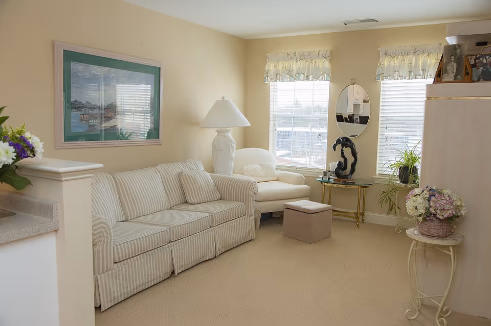 A cozy living room with a striped sofa, a white armchair, a floor lamp, and a small ottoman. There are two windows with white blinds and valances, a round glass table with a black abstract sculpture, and a small round side table with a basket of flowers. A framed picture hangs on the wall above the sofa.