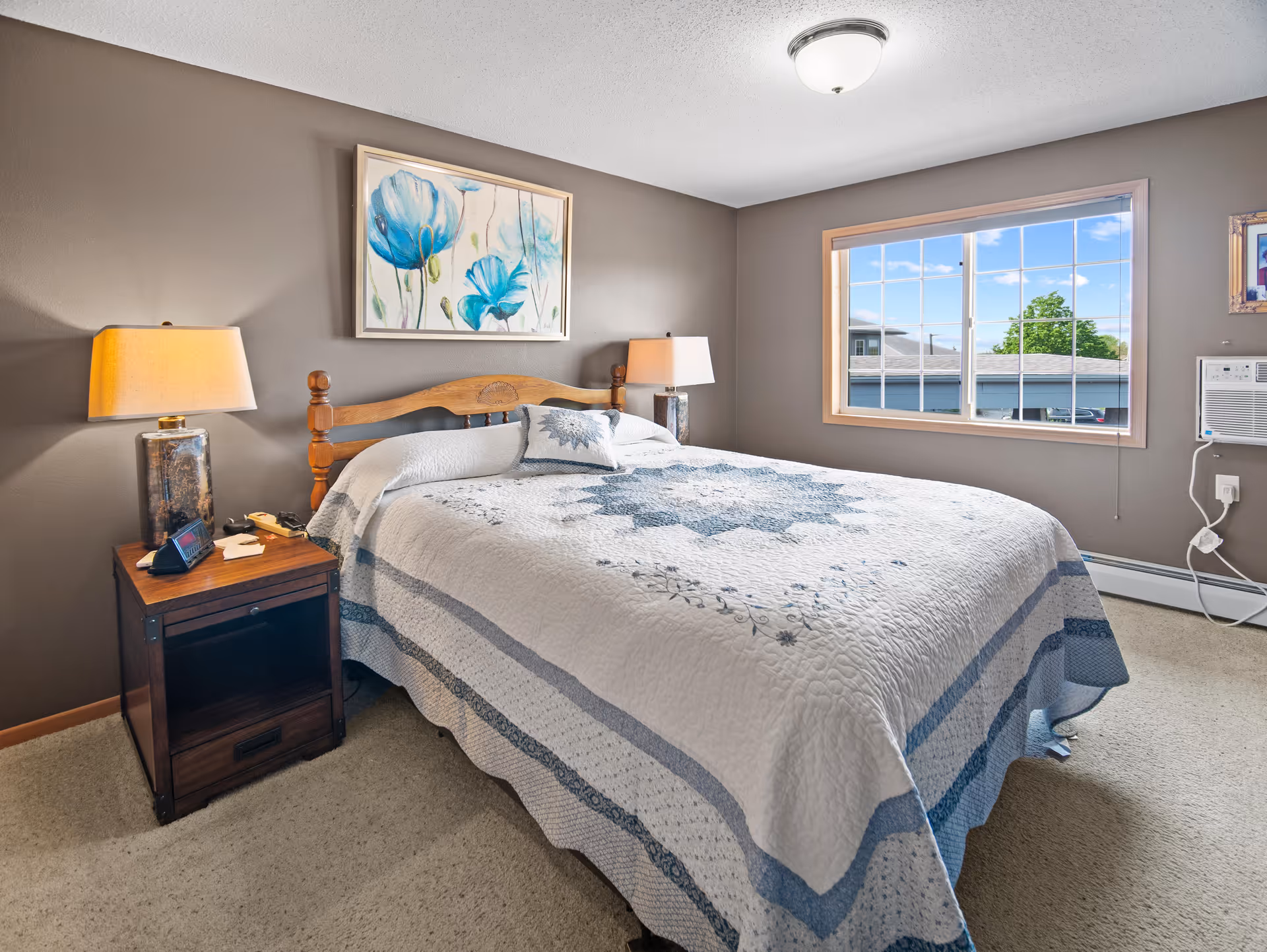 A cozy bedroom with a wooden bed frame and a white and blue quilted bedspread. There are two bedside tables with lamps on each side of the bed. A large window lets in natural light, showing a view of rooftops and a tree outside. The walls are painted gray and there is a floral painting above the bed.