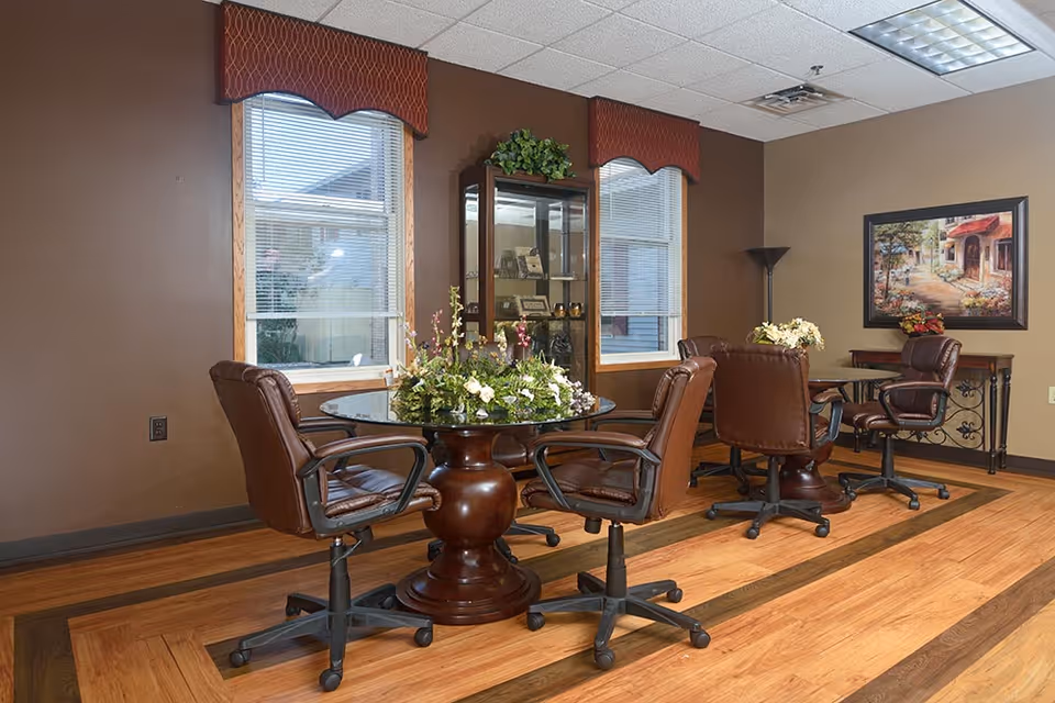 A cozy interior room with a round glass-top table surrounded by four brown leather swivel chairs on a wooden floor. The walls are painted brown and beige, with two windows featuring red valances. A glass display cabinet with decorative items stands between the windows, and a framed painting hangs on the beige wall. A floor lamp and a small table with a flower arrangement are also visible.