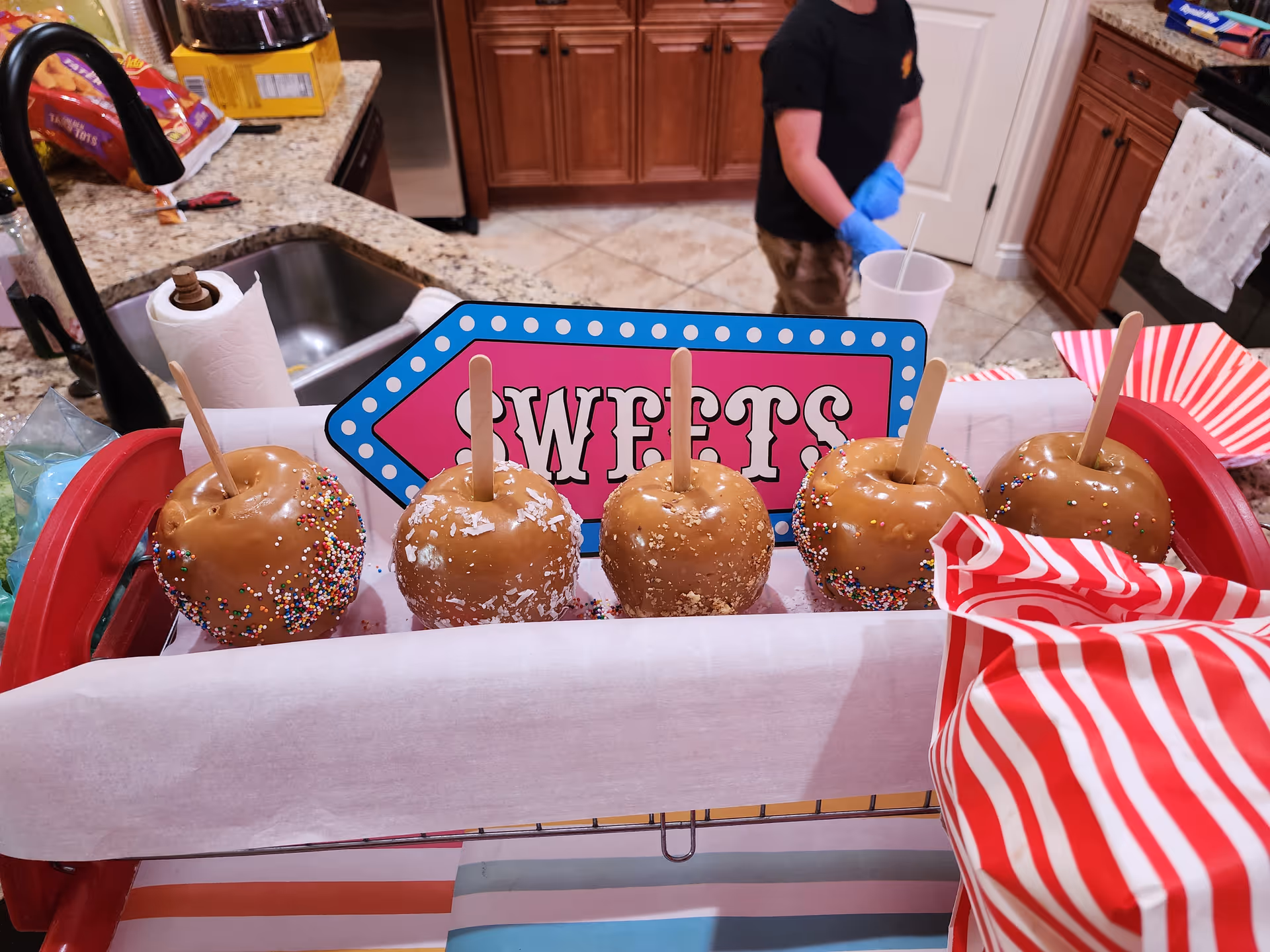 Caramel-coated apples with sprinkles arranged on a tray labeled 'Sweets' on a kitchen counter.
