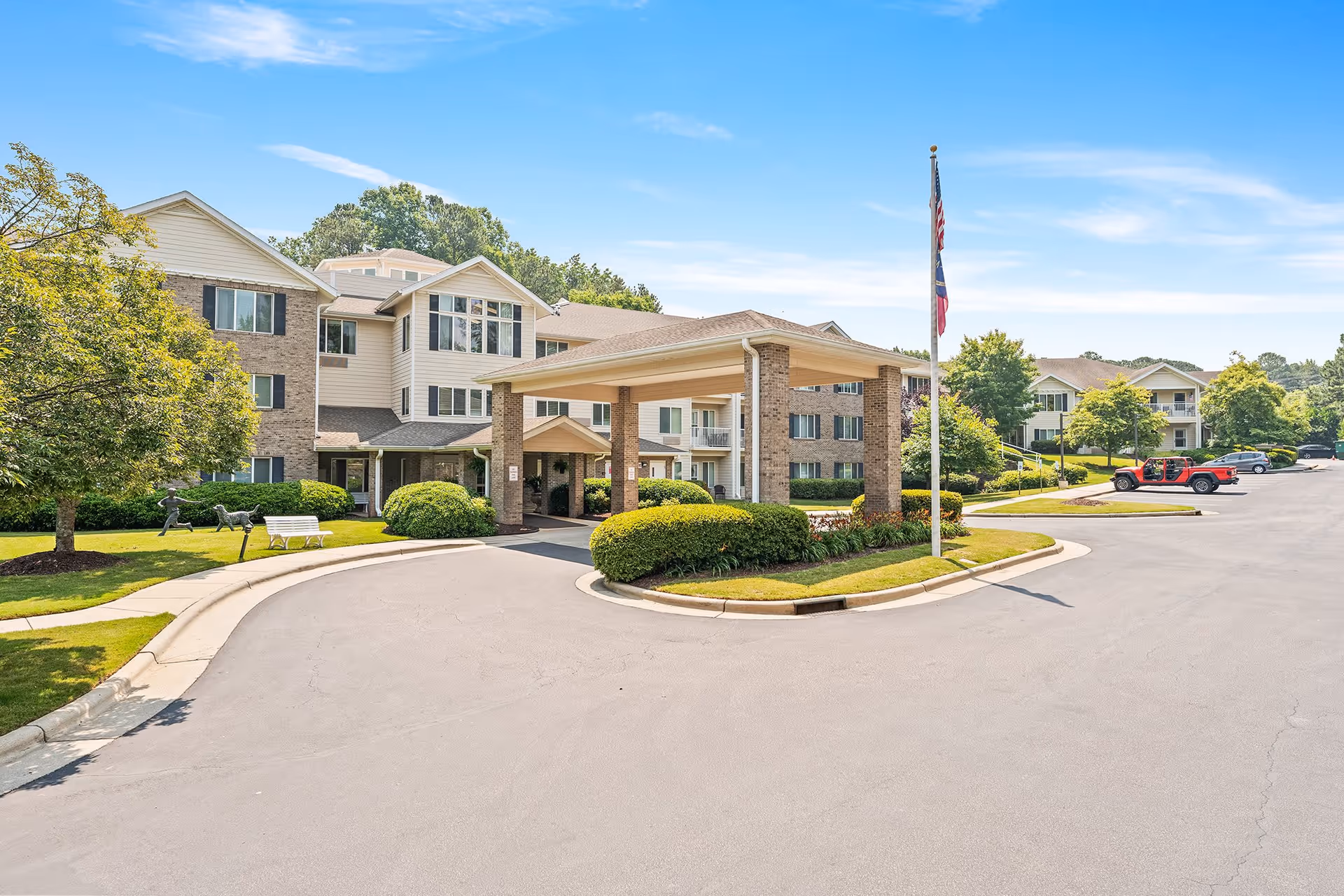 Exterior view of Jordan Oaks Gracious Retirement Living facility showing a multi-story building with a covered entrance, landscaped greenery, a flagpole with an American flag, and a parking area with several vehicles under a clear blue sky.
